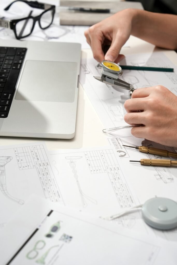 Close up of an eyewear designer using a dial caliper to measure a clear Vooglam frame on top of technical drawings beside a laptop and precision tools.