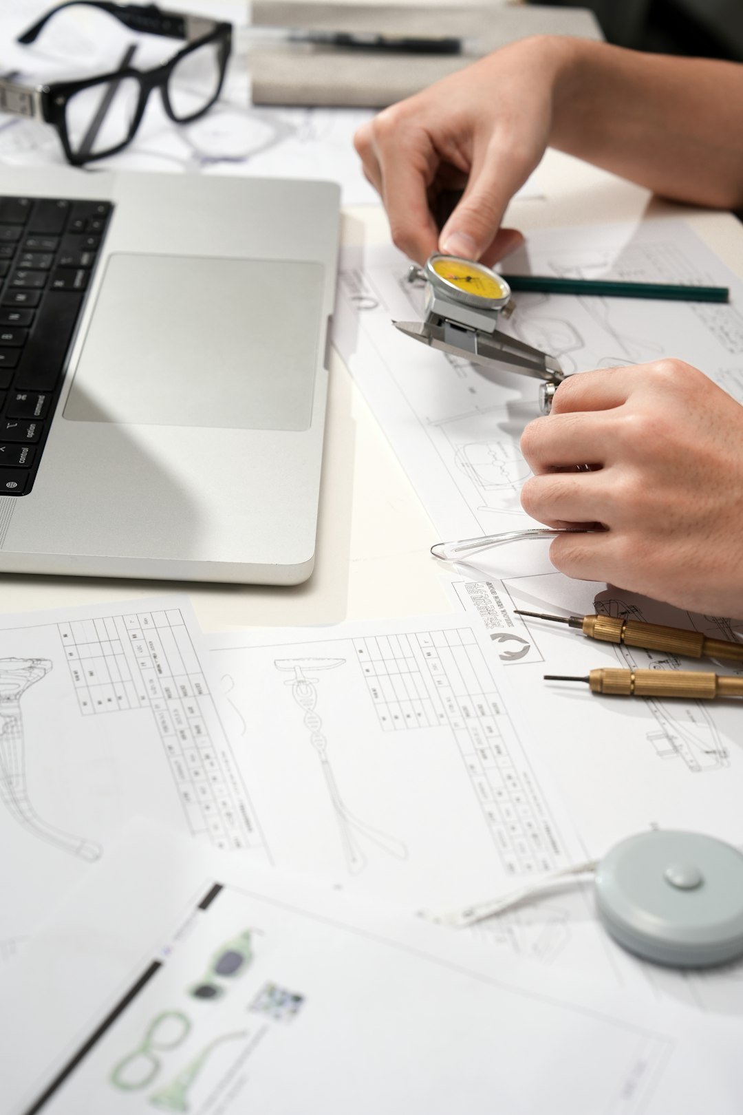 Close up of an eyewear designer using a dial caliper to measure a clear Vooglam frame on top of technical drawings beside a laptop and precision tools.