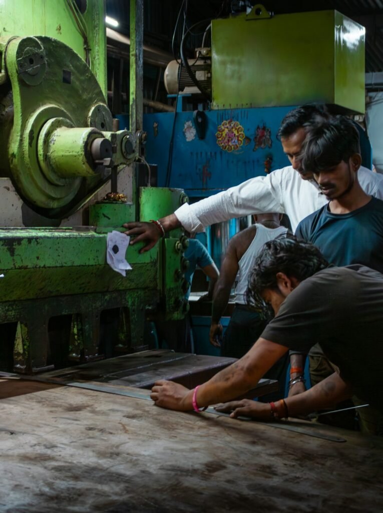 Inside a metal shearing, pressing, and cutting facility in Grant Road, India. Here a worker is setting the markings on a piece of metal before cutting it. The scene captures the collaborative nature of manual labor and industrial work in a small-scale setting, suitable for content on Indian manufacturing, working life, or traditional workshops.
